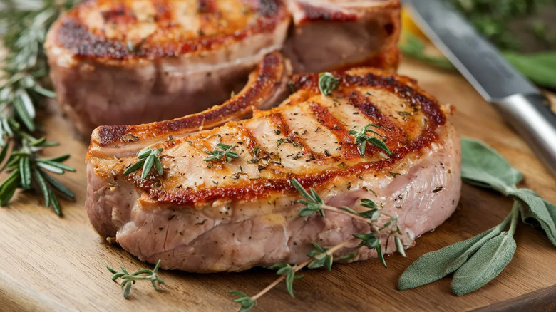 Thick-cut pork chops with grill marks sitting on wooden board with herb garnishes