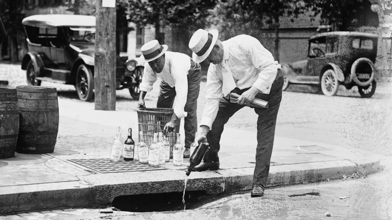 Two men in straw hats pour bottles of alcohol down a street drain during the Prohibition era, with several cars in the background