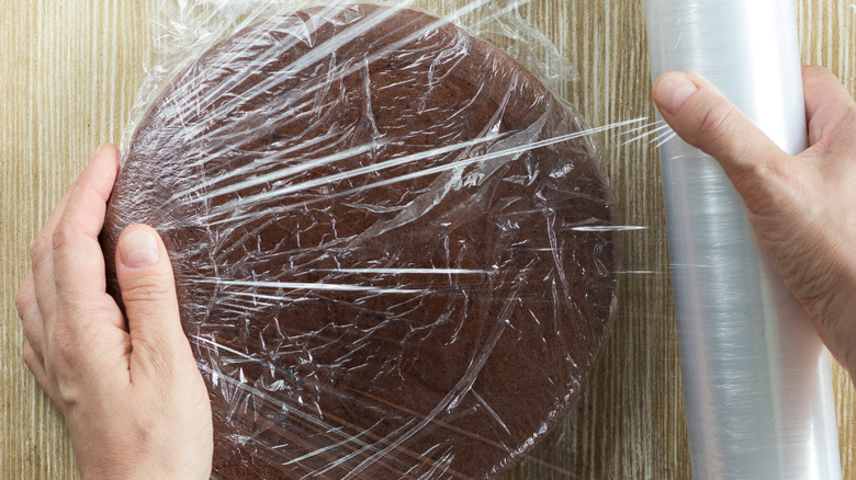 hands wrapping chocolate cake with plastic wrap for overnight storage