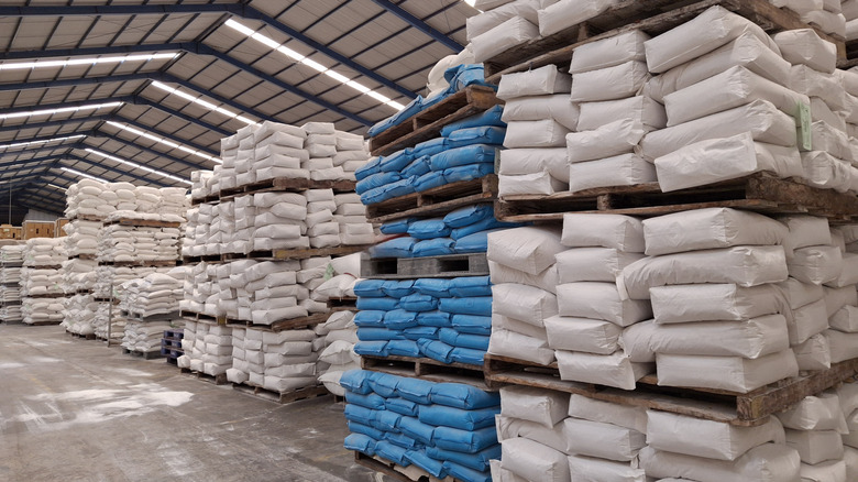 Pallets stacked high with sacks of basic ingredients, like flour and sugar, in a large, well-lit warehouse
