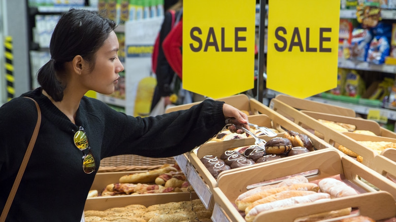 Young Asian woman using tongs to choose baked goods from a bulk bin, beneath a large yellow SALE sign