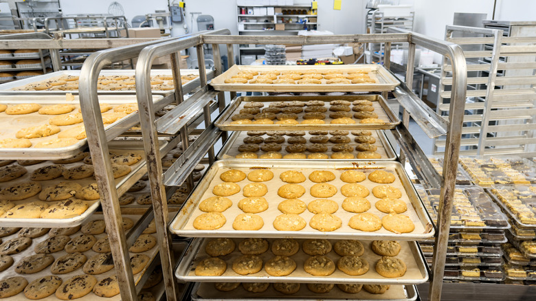 Multiple racks of cookies cooling in a large industrial bakery, with packages of other baked goods already packaged up for distribution to stores