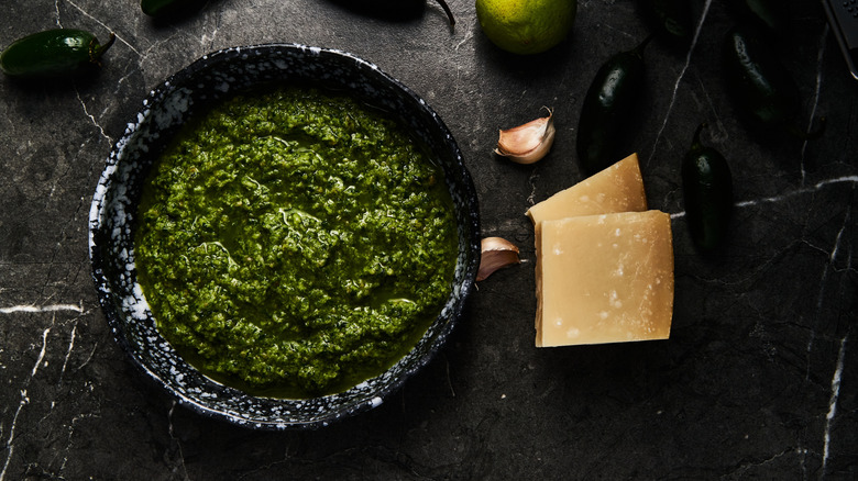 Bowl of jalapeño pesto next to wedges of Parmesan cheese, fresh jalapeños, and garlic cloves