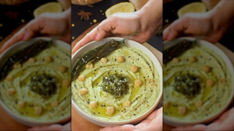 Hands holding a bowl of green jalapeño hummus, topped with roasted jalapeños and chickpeas