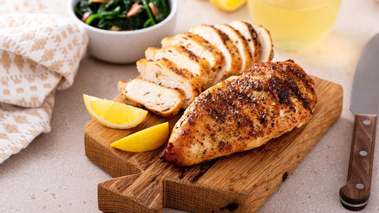 a whole grilled chicken breast next to a sliced one on a wooden cutting board with lemon wedges