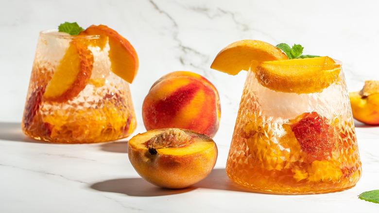 Two cocktails garnished with peach sliced on a white marble counter with peaches beside them