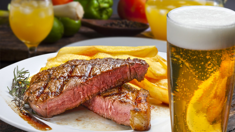 A steak cooked to medium and cut it in half, sitting on a plate with some french fries. There is a glass of beer in the foreground