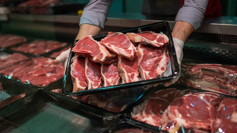 Butcher holding black tray of raw steak cuts in grocer's case.