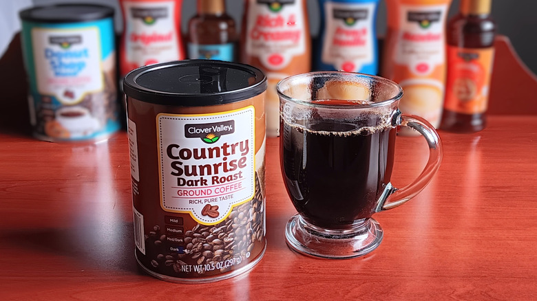 Can of Clover Valley Country Sunrise dark roast ground coffee next to a glass mug of black coffee.