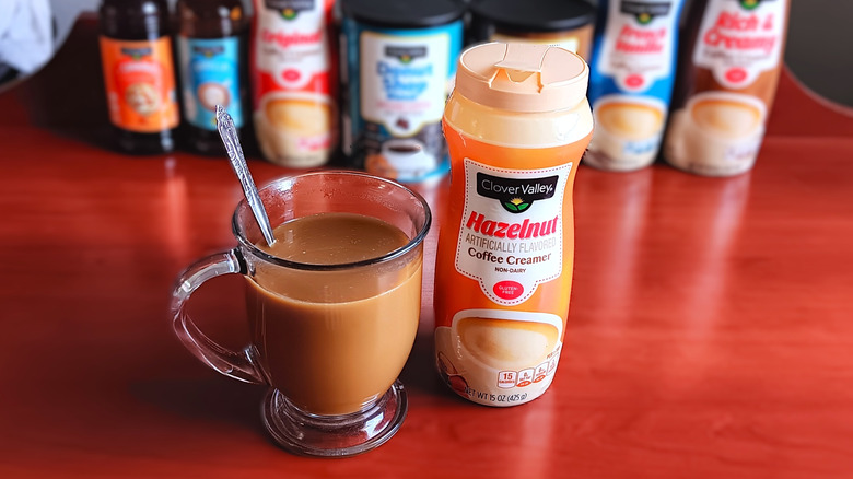 Glass mug of coffee and cream with spoon next to a container of Clover Valley hazelnut non-dairy coffee creamer.
