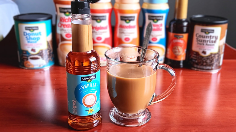 Bottle of Clover Valley vanilla syrup next to glass mug of coffee with cream and spoon.