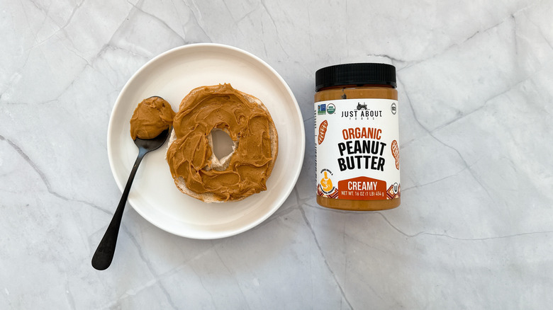 Peanut butter on a bagel and a jar of Just About peanut butter on a marble counter