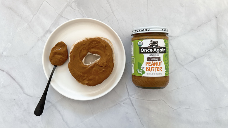 Half a bagel covered in peanut butter next to a spoon and a jar of Once Again peanut butter