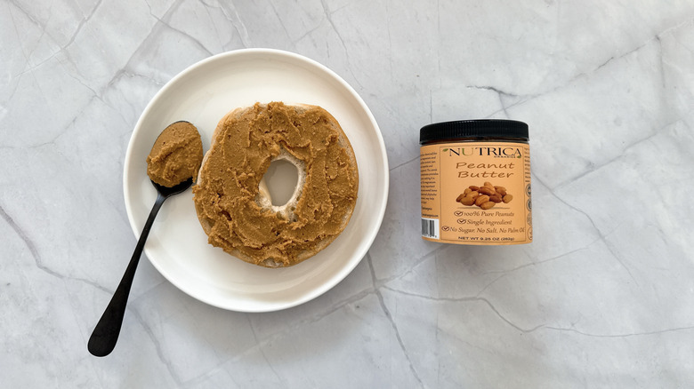 Half of bagel covered in peanut butter and jar of Nutrica peanut butter on the side