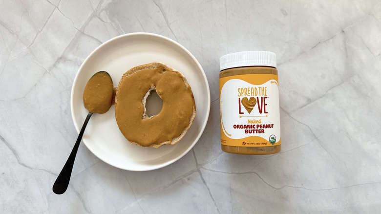 Bagel smeared with peanut butter on a white plate next to a jar of Spread the Love butter