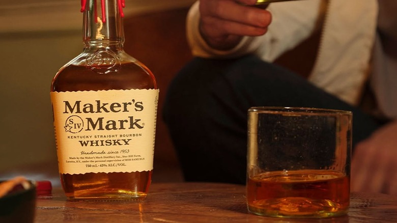 A bottle of Maker's Mark next to a filled glass and a person drinking in the background