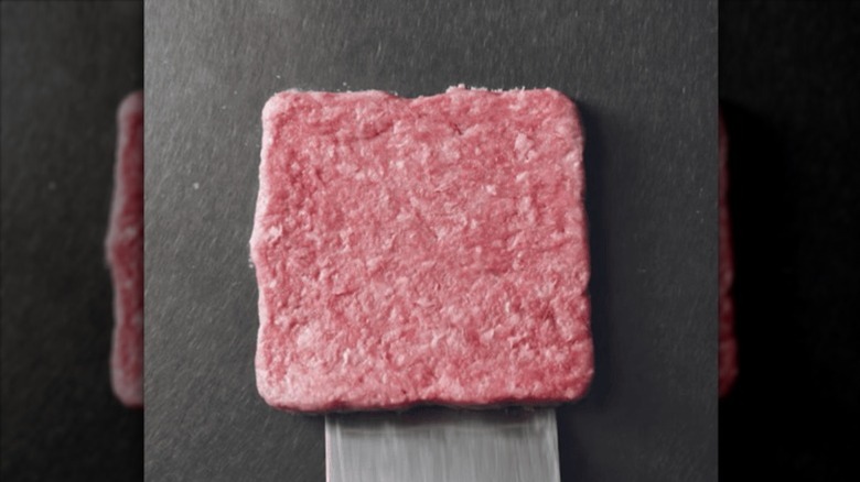 A metal spatula picking up a square hamburger patty