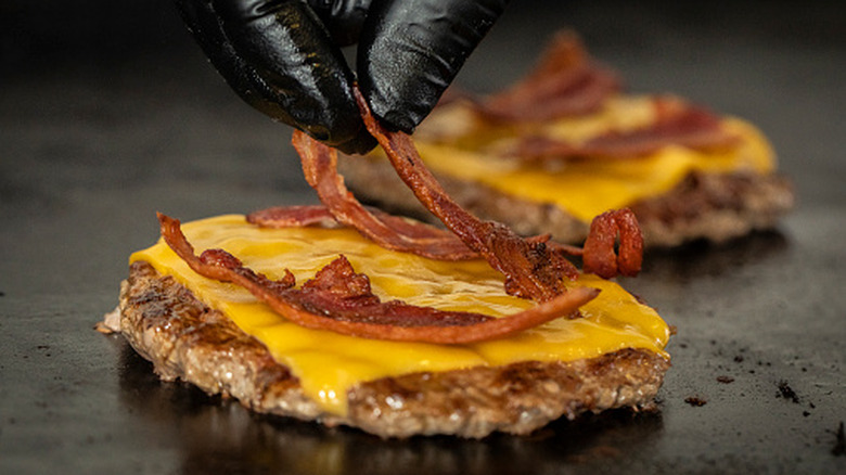 Gloved fingers placing strips of bacon onto a cheeseburger patty on the griddle with an identical burger patty in the background