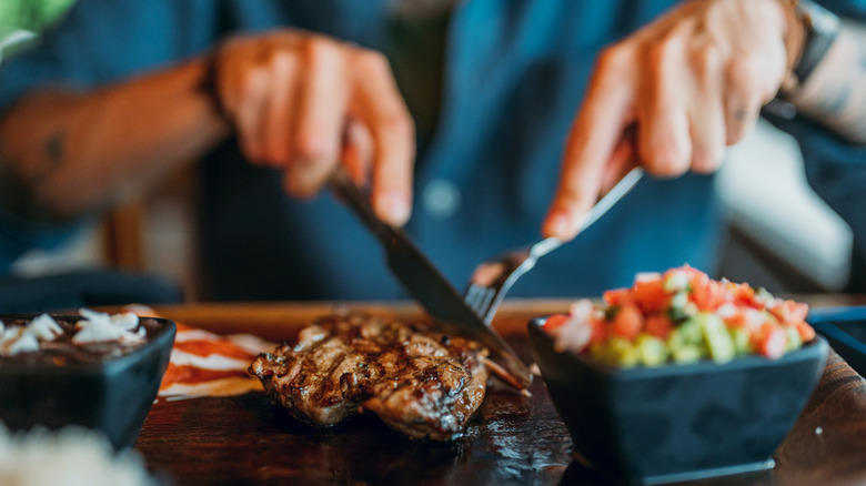 Hands holding knife and fork cutting steak on wooden board with blurred sides