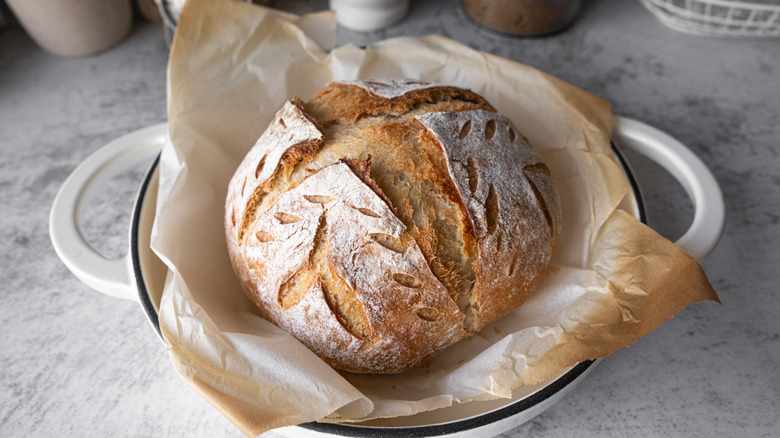 Round baked sourdough bread loaf sitting inside parchment paper-lined white enameled pot