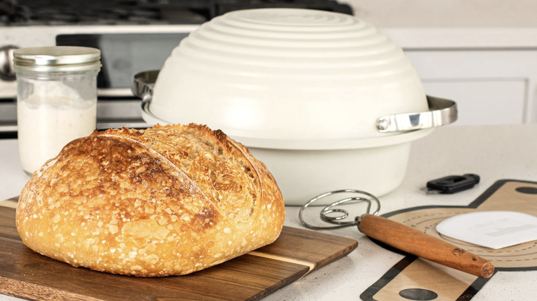 Loaf of sourdough bread on wooden board next to Nordic Ware brand baking tools including baking pan, dough hook, and silicone pan liner