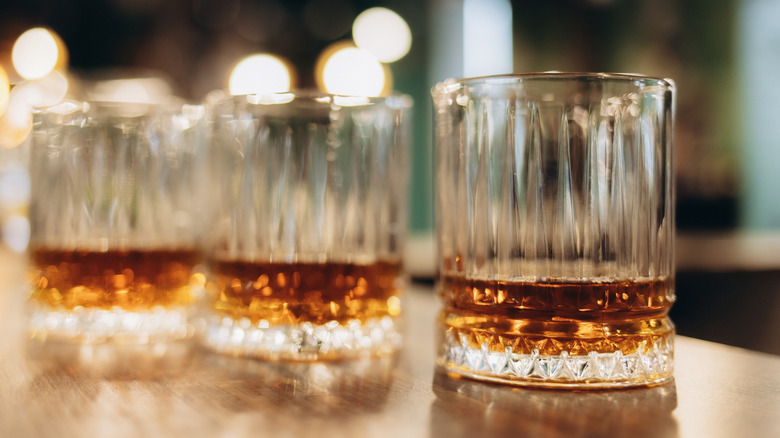 several glasses of whiskey on a bar with lights reflected in the background.