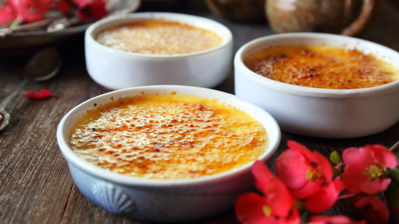 Three portions of crème brûlée in ceramic dishes on table alongside fresh flowers