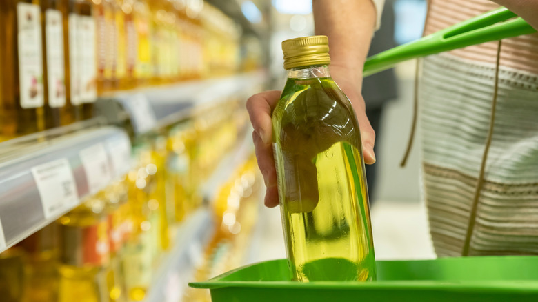 A person's hand places a bottle of olive oil in a basket while they stand at a supermarket shelf lined with olive oil bottles.