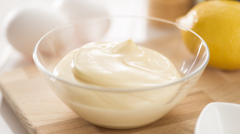 A bowl of homemade mayonnaise on a wooden cutting board with eggs and a lemon in the background