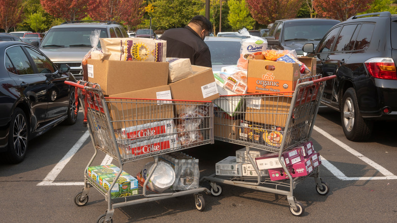 Customer in parking lot opening trunk of car with two crowded Costco carts full of goods in cardboard boxes nearby