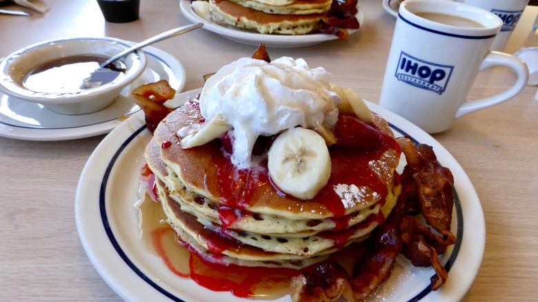 A plate of IHOP pancakes with syrup and jam rests on a tabletop with a coffee mug and bowl of syrup