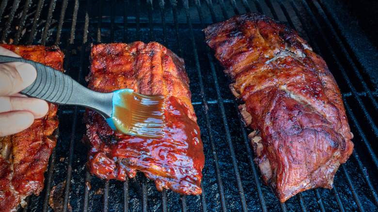 person's hand basting baby back ribs with a glaze on a grill