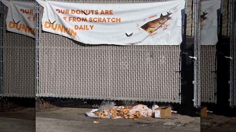 A pile of discarded Dunkin' food lying in front of a dumpster draped with a Dunkin' banner