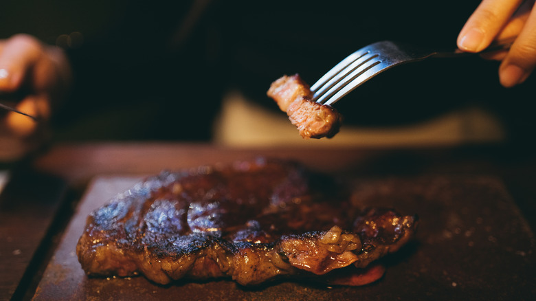 A person holds a bite of steak on a fork over a larger cut resting on a board