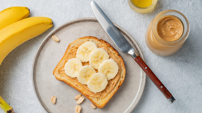 A plate containing a slice of banana peanut butter toast, with a bunch of bananas and a jar of peanut butter nearby