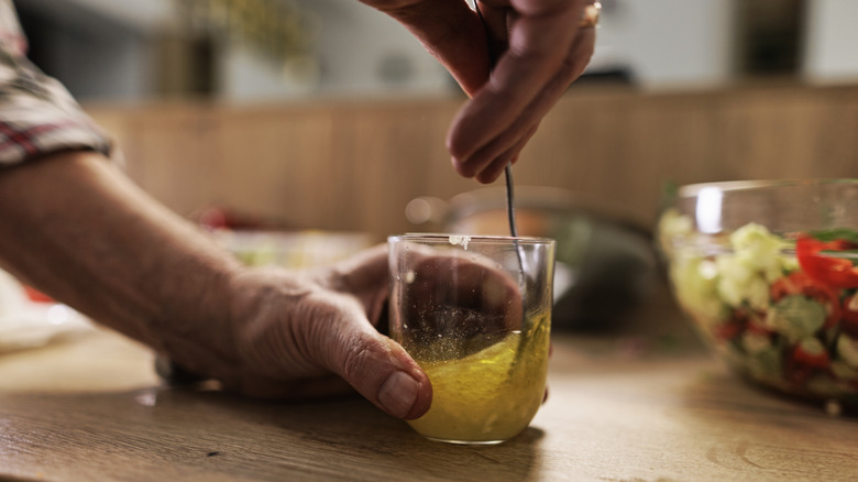 A hand steadies a glass cup full of oil and vinegar, while another hand stirs it with a fork