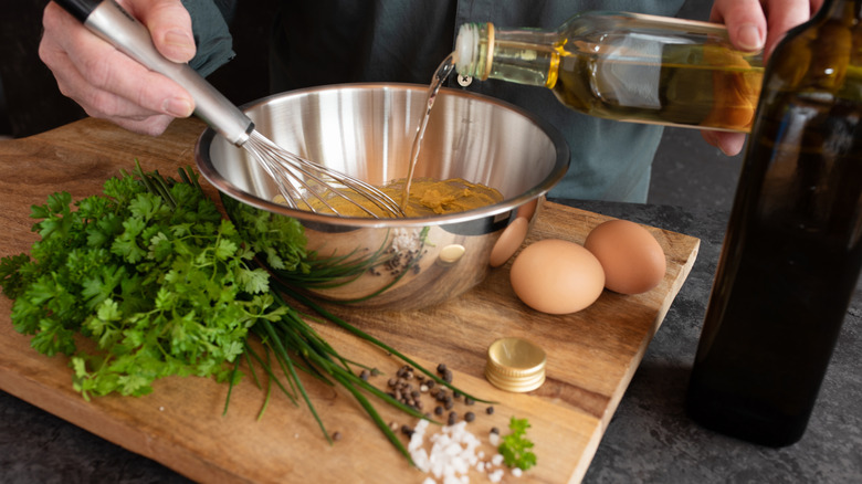 A person whisks oil into a bowl on a cutting board along with parsley, chives, salt, and pepper