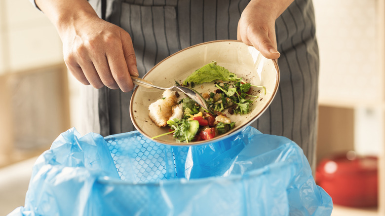 A chef in an apron scrapes leftover food into a garbage can