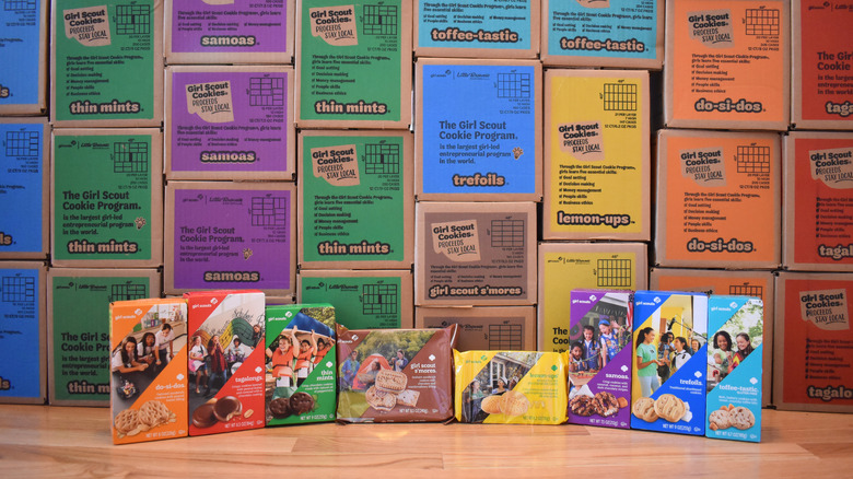 Boxes of Girl Scout Cookies lined up on a table in front of bulk cookie boxes in the background