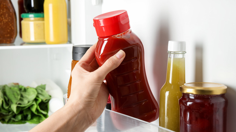 A woman's hand reaching for an unlabeled bottle of ketchup in the fridge