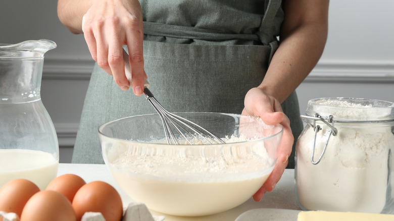 Woman's hands mixing batter at counter next to jar of flour, milk jug, and eggs