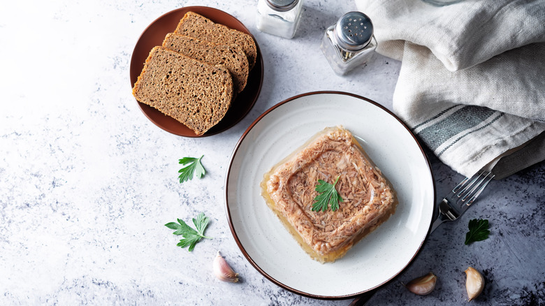 meat aspic on a white plate next to bread on a red plate tablecloth and seasoning