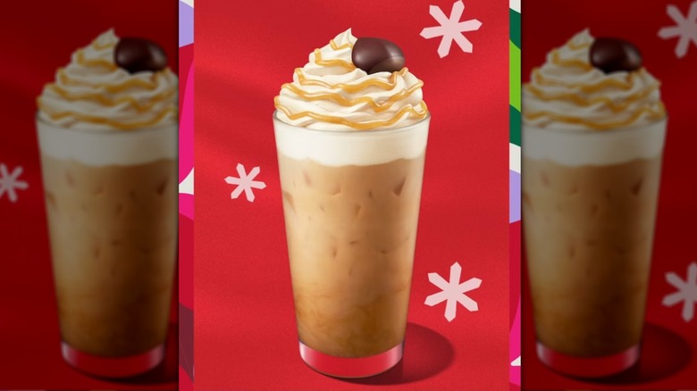 A tall glass is filled with a creamy caramel brown liquid over ice and topped with whipped cream, a caramel colored sauce is drizzled on top, and there is a chestnut-shaped chocolate on top. The background is bright red and adorned with three white snowflakes