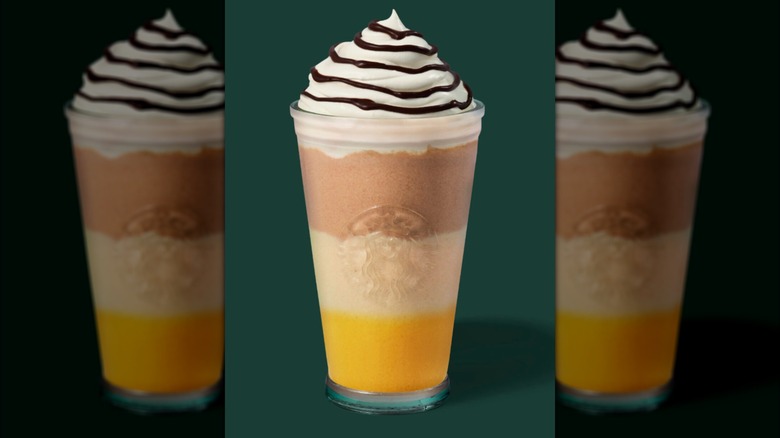 A Starbucks-branded tumbler is filled with a bright yellow liquid at the bottom, the next layer is a creamy white color, and the third layer on top is a darker coffee or chocolate brown. The beverage features whipped cream on top and a drizzle of a dark chocolate or mocha sauce