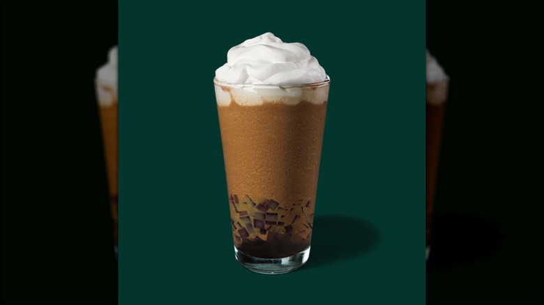 A tall clear glass is filled with coffee jelly squares on the bottom, iced milk tea, and a vanilla cold foam on top