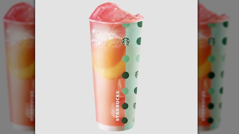 A reusable Starbucks cup is filled with a bright pink liquid and featured with two designs. One vertical half of the cup is mint green and features circular dot pattern with various shades of green reminiscent of the Starbucks logo itself, which is mixed in with the other green dots. The other half of the cup is a grapefruit-red color and features an image of a yellow peach surrounded by carbonated bubbles.