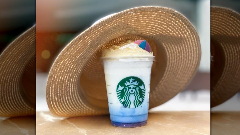 A Starbucks branded cup features a blue liquid at the bottom and gradually whitens to a creamy white liquid near the top. Whipped cream is added with an umbrella-shaped cookie and graham cracker crumbs on top. This drink is wearing a light brown straw beach hat.