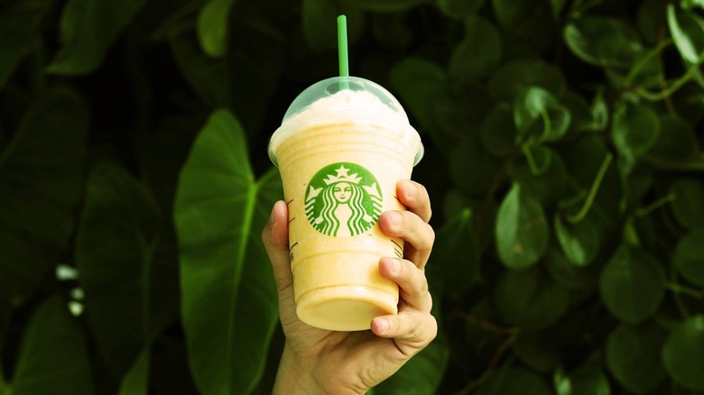 A hand holds up a Starbucks-branded cup that is filled with a yellow substance, whipped cream, and a green straw in front of big green leaves seemingly from some kind of plant or tree.