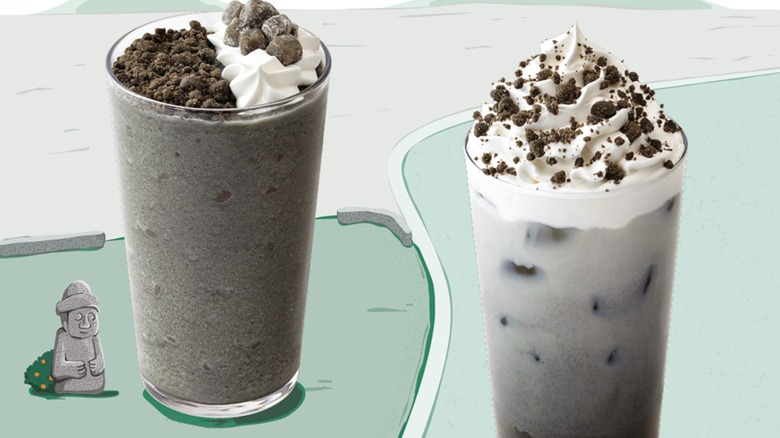A mini gray statue cartoon sits next to a tall glass filled with a dark gray, almost black frozen liquid that is topped with a black crumb. A similar dark gray drink with a creamier milky consistency stands next the first glass