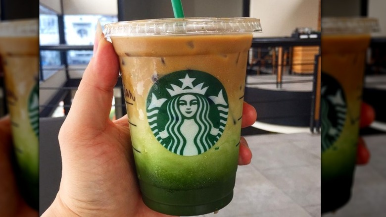 A clear Starbucks-branded cup is filled with a green matcha powder on the bottom. There's a milky white creamy layer on top of the green matcha, and a caramel brown colored coffee or espresso layer on top. The cup has a lid and a green straw in it. A hand is holding the cup up to the camera and some items in the background are visible like tables and stairs.